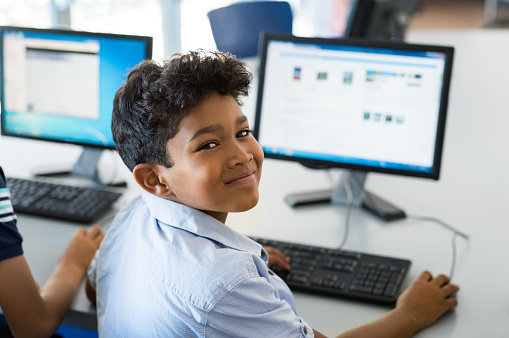 Young happy schoolboy using computer to search internet. Arab child learning to use computer at elementary school. Portrait of smiling middle eastern kid looking at camera while surfing the net in school library.
